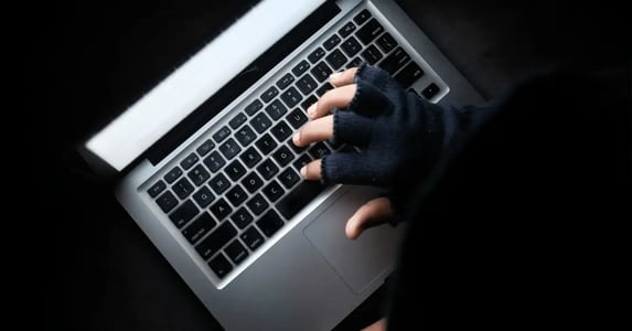 An overhead view of a shrouded figure in a dark room typing on a laptop computer.